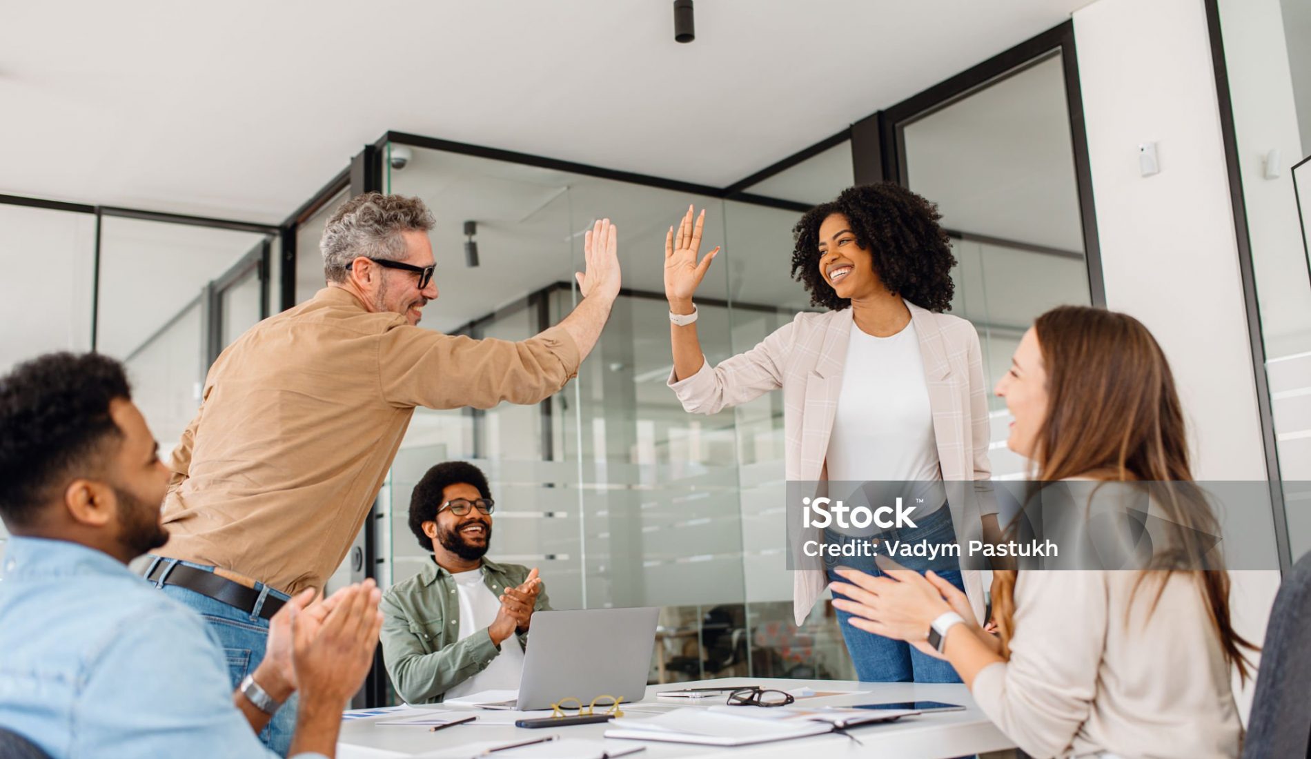 A diverse team of professionals celebrates a workplace achievement with a high-five in a modern office. The moment captures motivation, cooperation, and a positive business culture focused on teamwork